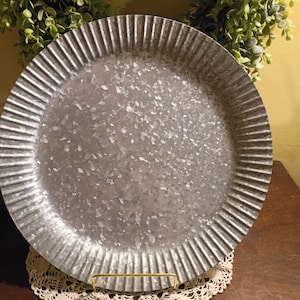 May include: A galvanized metal tray with a fluted rim. The tray is round and sits on a cream-colored lace doily. The tray is displayed on a gold-colored metal stand. A green wreath is in the background.