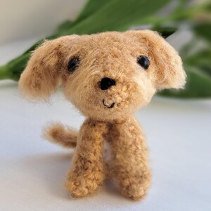May include: A small, handmade, tan-coloured dog figurine with black eyes and a smiling mouth. The plush toy has floppy ears and a small tail. The dog is standing on a white surface, with green foliage in the background.
