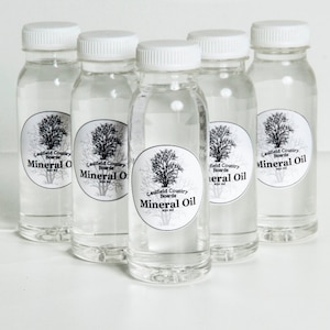 Può includere: Cinque bottiglie di vetro trasparente con tappi bianchi ed etichette nere. Ogni etichetta presenta un disegno di un albero e il testo "Caulfield Country Boards Mineral Oil".