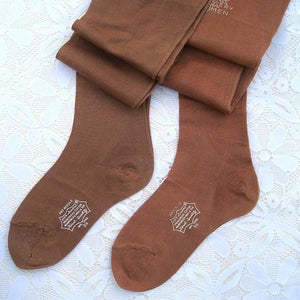 May include: Two pairs of brown hosiery, folded and arranged on a white lace surface. The hosiery features white text and a logo that reads "Silver Seal Hosiery Women" and "First Quality". The socks are made of a soft material.