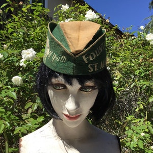 May include: A vintage green and tan hat with the words "Fountain St. Aubin" and "Fountain of Youth" embroidered on it. The hat is displayed on a mannequin head with black hair and green eyes, set against a backdrop of white roses and greenery.