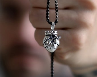 May include: A silver heart pendant with a crown on top hangs from a black chain. The pendant is being held by a hand.