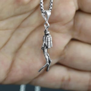 May include: A silver-colored pendant necklace featuring a detailed miniature diver. The diver is suspended from a chain. The pendant depicts a diver with scuba gear, legs extended, and arms at their sides. The necklace is held in a hand.