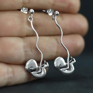 May include: Silver-toned earrings featuring a stylized fetus design. The earrings have a post-back closure and a curved, dangling design. The fetus is depicted in a curled position, with a smooth, rounded form. The earrings are held in a hand against a dark background.