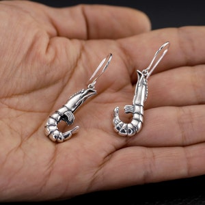 Può includere: Un paio di orecchini a forma di gambero in argento. I gamberi sono dettagliati e realistici, con le chele ricurve. Gli orecchini sono appesi a ganci d'argento.