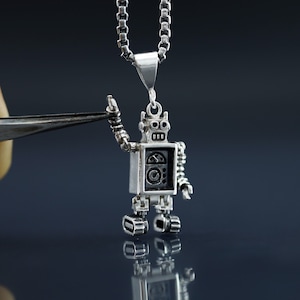 Könnte beinhalten: Eine silberne Roboter-Anhängerkette mit einer Kette. Der Roboter steht mit erhobenen Armen. Der Roboter hat ein schwarzes Gesicht mit zwei Augen und einem Mund.