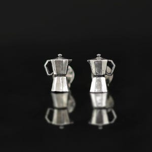 Könnte beinhalten: Ein Paar silberfarbene Manschettenknöpfe mit Kaffeekannen-Motiv. Die Manschettenknöpfe sind mit einem detaillierten Design einer klassischen italienischen Espressokanne versehen. Der Hintergrund ist eine dunkle, reflektierende Oberfläche.