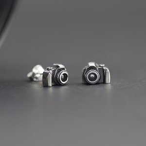 Puede incluir: Un par de pendientes de plata en forma de cámara. Los pendientes son pequeños y detallados, con un diseño de cámara realista.