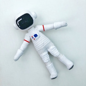 May include: White crocheted astronaut doll with a black visor and red and blue accents. The doll is lying on its back with its arms outstretched.