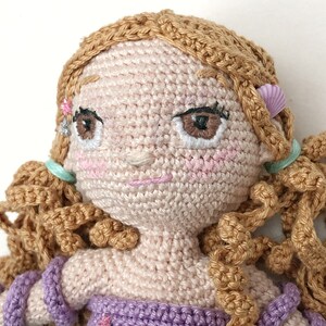 May include: A crocheted doll with brown hair, pink cheeks, and embroidered eyes. The doll is wearing a purple dress and has a pink seashell on its hair.