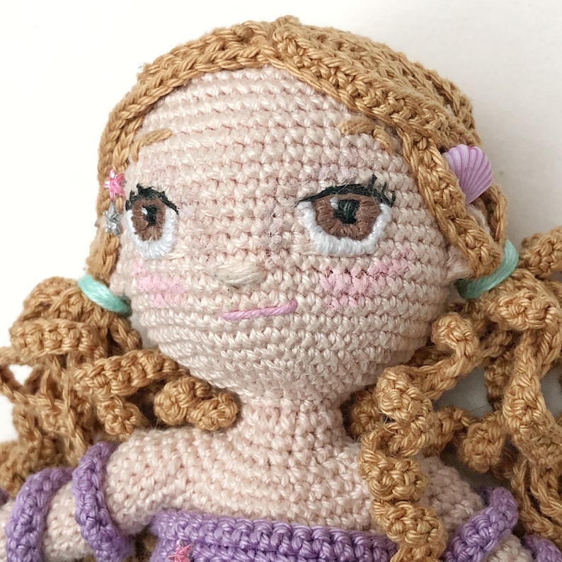 May include: A crocheted doll with brown hair, pink cheeks, and embroidered eyes. The doll is wearing a purple dress and has a pink seashell on its hair.