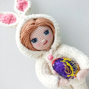 May include: A white crocheted bunny doll with blue eyes and pink ears, wearing a white hooded sweater and holding a purple and gold chocolate egg.