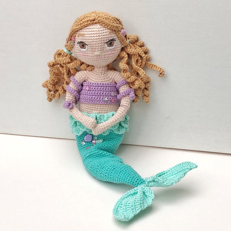 May include: A crocheted mermaid doll with long brown hair, a purple top, and a green tail. The doll has a sweet face with pink cheeks and blue eyes.