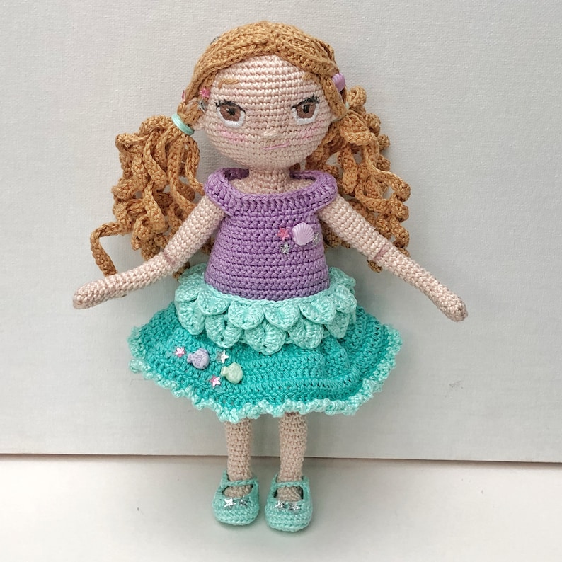 May include: Crocheted doll with long brown hair, wearing a purple bodice and a teal skirt with a scalloped edge. The doll has a sweet expression and is wearing teal shoes.