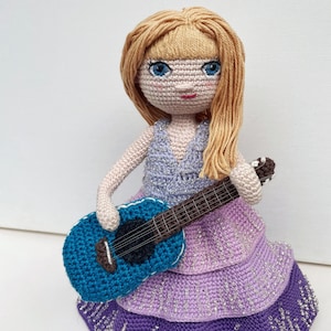 May include: A crocheted doll with long blonde hair wearing a purple dress and holding a blue guitar.