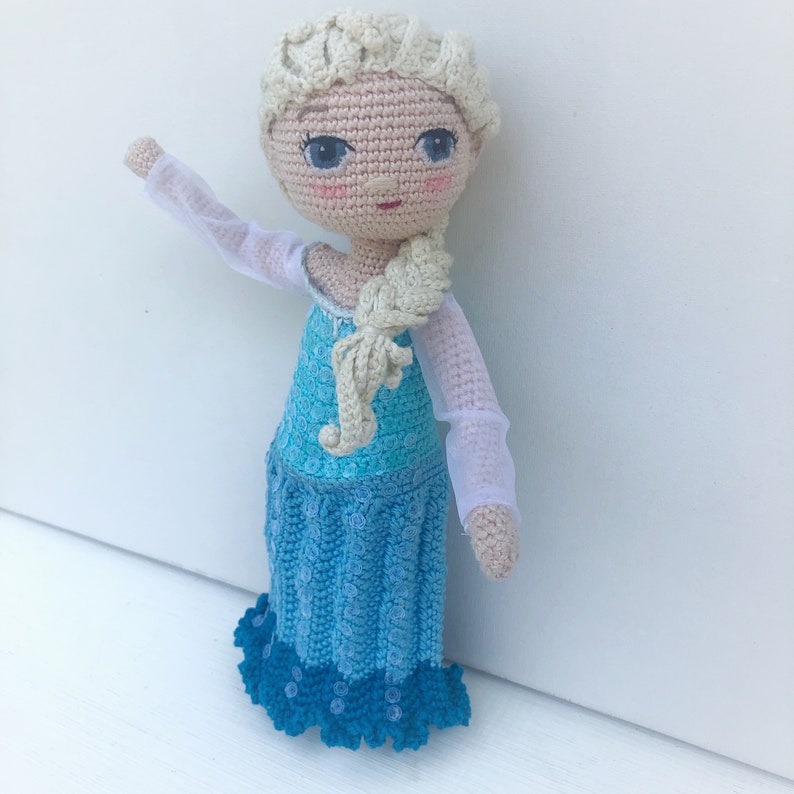 Pu&ograve; includere: Una bambola all'uncinetto con una treccia bionda, vestita con un abito blu e bianco con un'applicazione di pizzo bianco. La bambola ha gli occhi azzurri e un blush rosa.