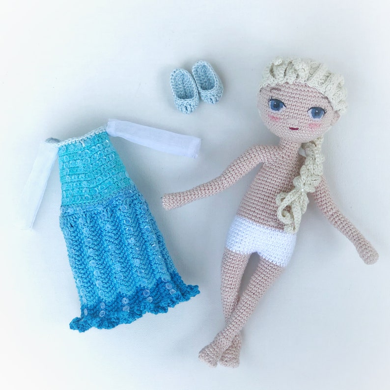 Pu&ograve; includere: Una bambola all'uncinetto con i capelli biondi che indossa un vestito bianco all'uncinetto e un vestito blu all'uncinetto. La bambola &egrave; sdraiata su una superficie bianca con due scarpe blu all'uncinetto.