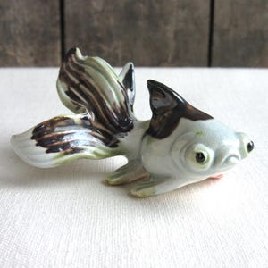 May include: A small ceramic figurine of a black and white goldfish with green and brown markings on its tail fins. The goldfish has large, round eyes and a slightly open mouth.