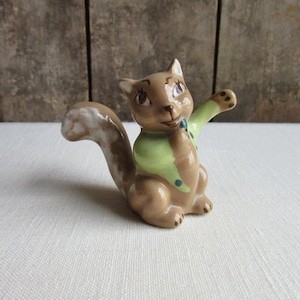 May include: A small ceramic squirrel figurine with a brown and white tail, wearing a green shirt with blue polka dots and a white collar. The squirrel is sitting up with its right paw raised.