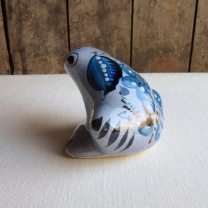 May include: A blue and white ceramic frog figurine with a floral design. The frog is sitting on its hind legs with its front legs tucked in. The frog is painted with blue, white, and brown details. The frog has a small inscription on its side that reads "Tlaquepaque Mexico".