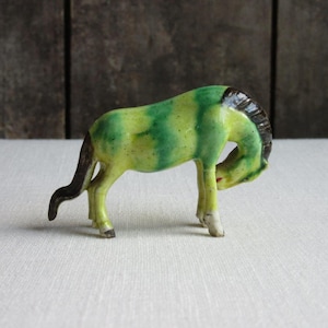 May include: A small ceramic horse figurine with green and yellow glaze. The horse is in a bowing position with its head down.