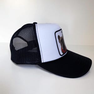 May include: Black and white trucker cap with a mesh back. The front panel is white with a patch featuring a French Bulldog wearing a red collar. The peak is black.