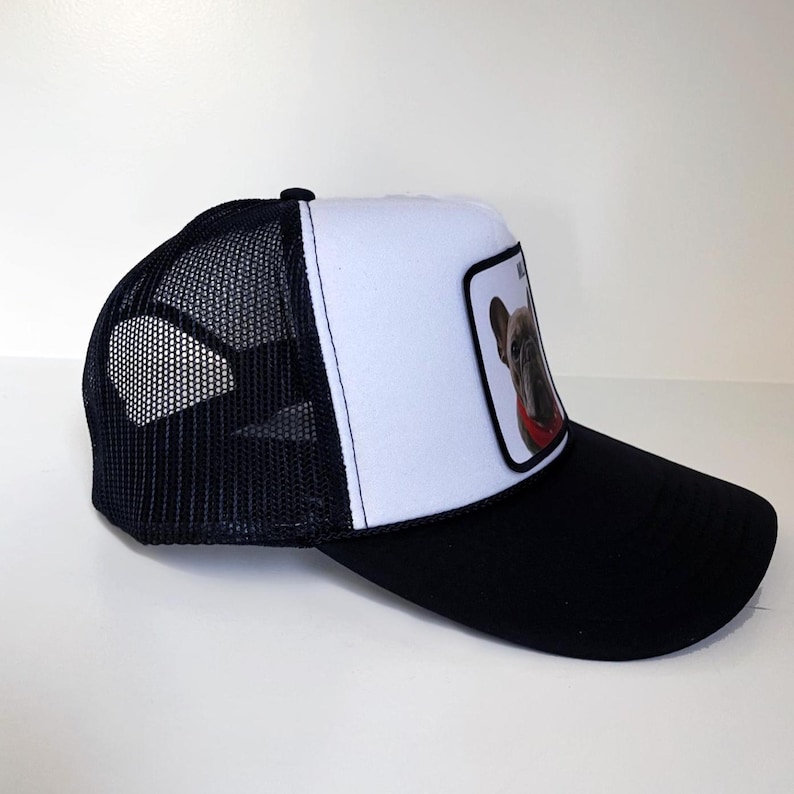 May include: Black and white trucker cap with a mesh back. The front panel is white with a patch featuring a French Bulldog wearing a red collar. The peak is black.