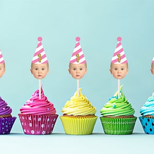 Op de afbeelding: Vijf cupcakes met roze en wit gestreepte feestmutsen met het nummer "1" erop. Elke cupcake heeft een andere kleur glazuur: paars, roze, geel, groen en blauw.