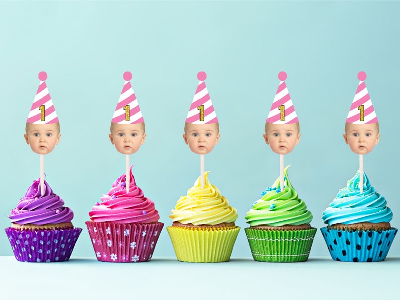 Op de afbeelding: Vijf cupcakes met roze en wit gestreepte feestmutsen met het nummer "1" erop. Elke cupcake heeft een andere kleur glazuur: paars, roze, geel, groen en blauw.