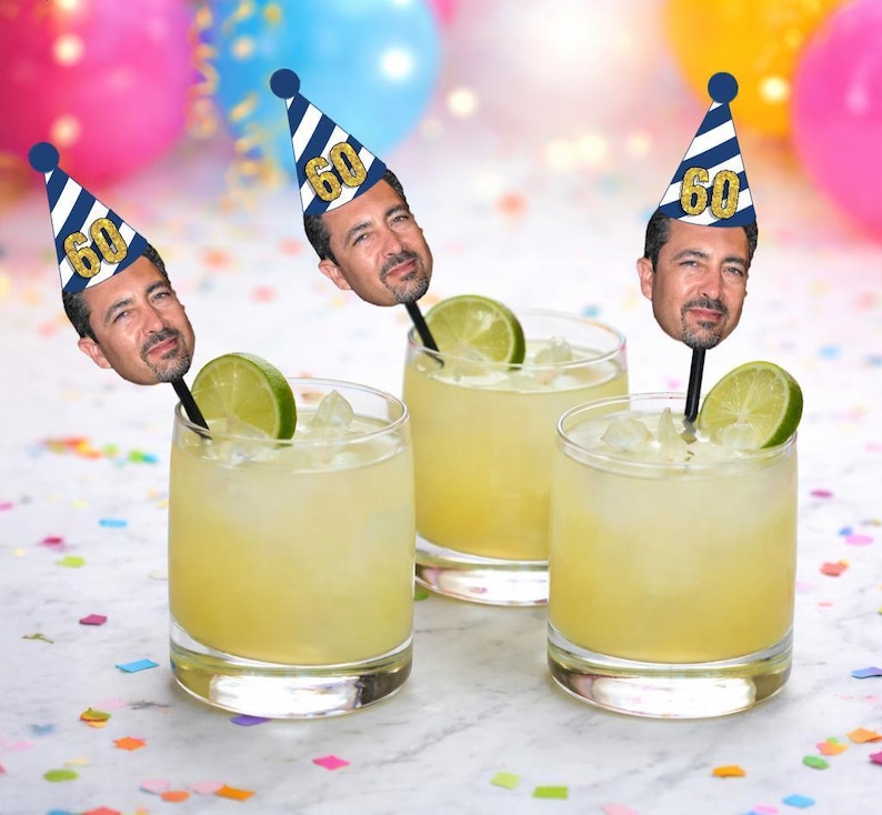 May include: Three cocktails in clear glasses with lime wedges and ice. Each drink has a black straw topped with a party hat with the number 60 and a face. Colorful balloons and confetti are in the background.