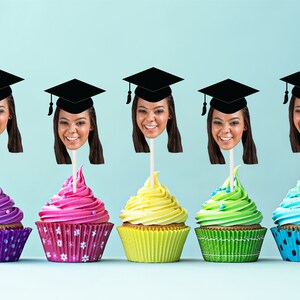 May include: Five cupcakes decorated with colorful frosting and topped with graduation caps and faces. The cupcakes are arranged in a row on a light blue background.