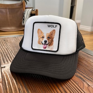 May include: A black and white trucker hat with a square patch showing a corgi dog portrait and the word "WOLF". The hat has a black peak and a mesh back.