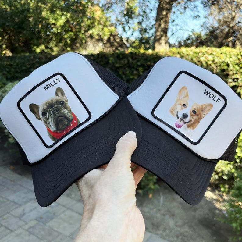 May include: Two black and white trucker caps. One cap features a French bulldog with the name "MILLY" above the image. The other cap has a corgi dog image with the name "WOLF". Both caps have a black mesh back and a white front panel.