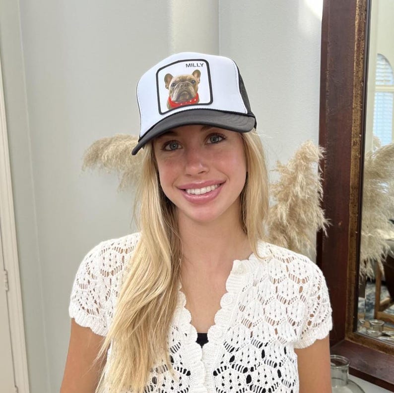 May include: A white and black trucker hat with a square patch showing a bulldog illustration and the name "MILLY". The hat has a black peak and mesh back.