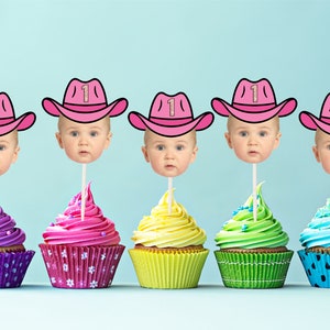 May include: Five cupcakes decorated with pink, purple, yellow, green, and blue frosting. Each cupcake has a pink cowboy hat with a gold glitter number one on top. The hats are attached to sticks and have a baby's face on each stick.