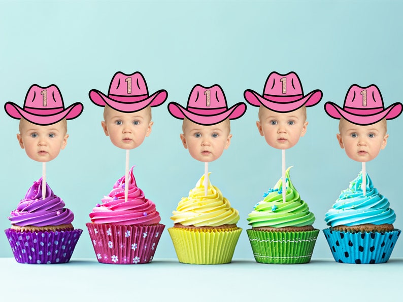 May include: Five cupcakes decorated with pink, purple, yellow, green, and blue frosting. Each cupcake has a pink cowboy hat with a gold glitter number one on top. The hats are attached to sticks and have a baby's face on each stick.