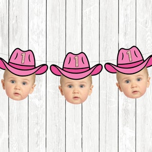 May include: A pink and white banner with four baby faces wearing pink cowboy hats with the number "1" on them. The banner is hanging on a white wood background.