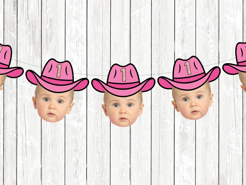May include: A pink and white banner with four baby faces wearing pink cowboy hats with the number "1" on them. The banner is hanging on a white wood background.