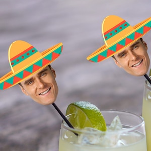 May include: Two yellow and green sombreros with a person's face on the top of each. The sombreros are on sticks that are in two glasses of a light yellow liquid. The glasses are on a wooden surface.