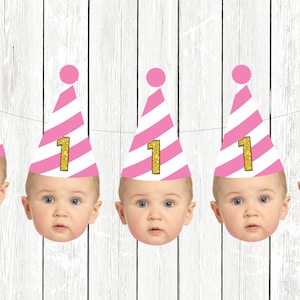 Op de afbeelding: Een roze-wit gestreepte feestbanner met vijf babygezichten die roze-witte feestmutsen dragen met gouden glitter "1"s.