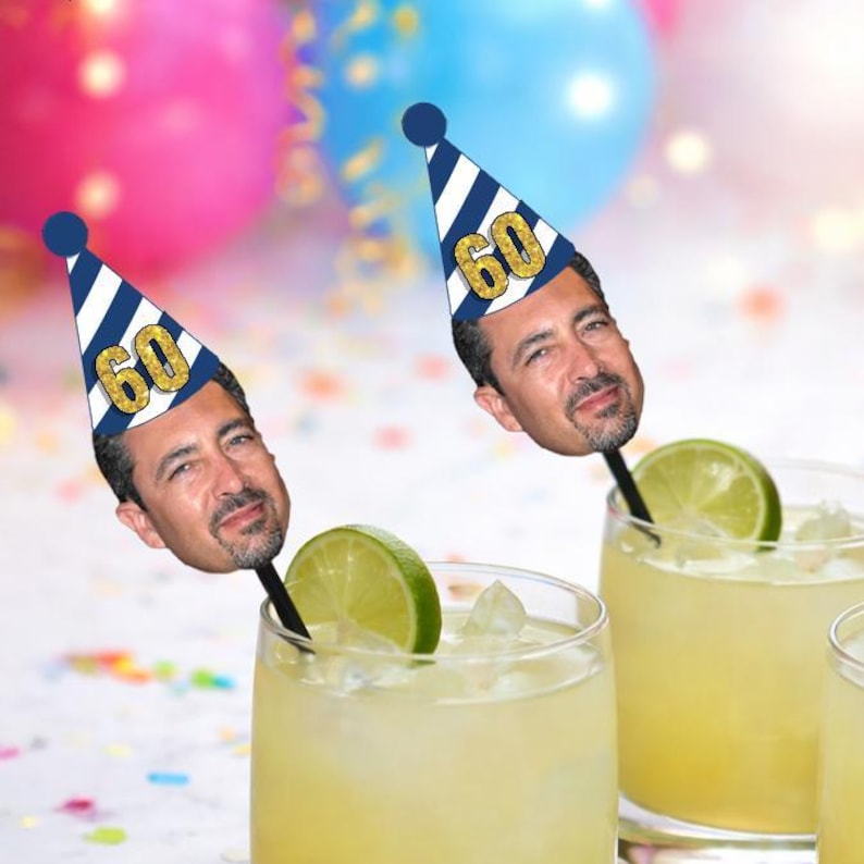 May include: Two cocktail glasses with lime wedges and drink stirrers featuring a man's face wearing a party hat with the number 60. The party hats are navy blue and white striped with a gold number 60. Colorful balloons are in the background.