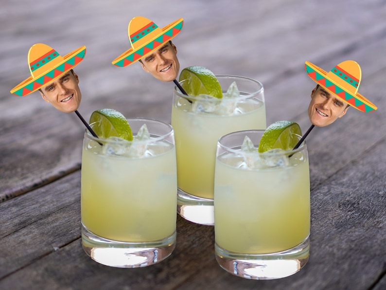 May include: Three clear glasses filled with a yellow liquid and ice cubes. Each glass has a lime wedge and a cocktail stick with a small sombrero and a man's face on top.