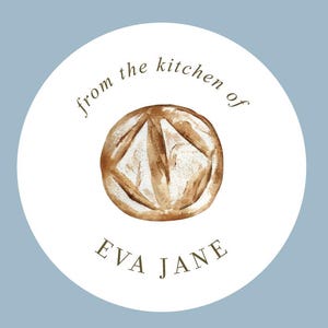 May include: A circular white sticker featuring a brown watercolor illustration of a round loaf of bread in the center. The text "from the kitchen of" is arched above the bread, and the name "EVA JANE" is printed in dark brown below the bread.