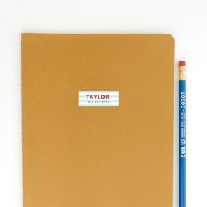May include: A brown notebook with a white and blue striped label that says "TAYLOR 212 355 8732" on it. A blue pencil is next to the notebook.