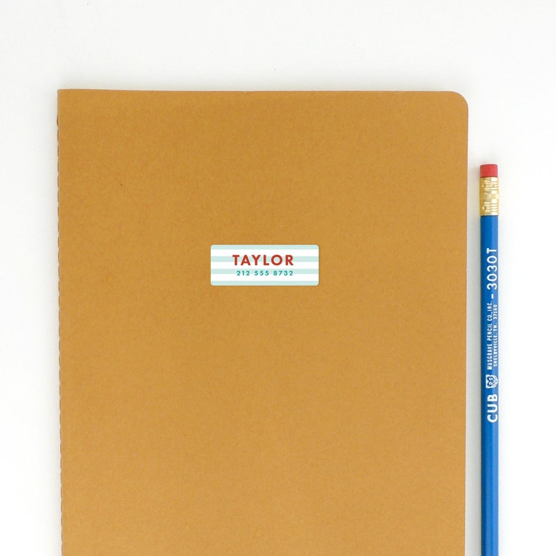 May include: A brown notebook with a white and blue striped label that says "TAYLOR 212 355 8732" on it. A blue pencil is next to the notebook.