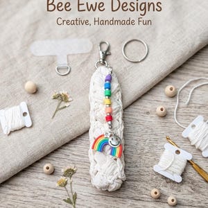 May include: A white crocheted keychain featuring a silver lobster clasp, a vertical row of multi-colored beads, and a dangling charm with a rainbow and a smiley face. The text Bee Ewe Designs Creative, Handmade Fun is visible at the top.
