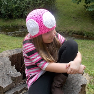 May include: A pink and white crochet mushroom hat. The hat has a rounded top with two large white circles on the front. The person wearing the hat is also wearing a pink, black, and white striped shirt and black pants.