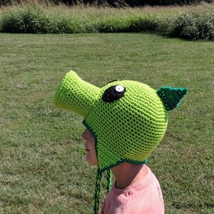 Puede incluir: Un gorro de crochet verde y negro con forma de monstruo vegetal con un hocico largo y un solo ojo negro. El gorro tiene un borla verde y negro que cuelga del extremo del hocico.