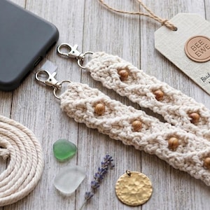 May include: Cream-colored macrame phone lanyard with wooden beads and silver-tone clasps. The lanyard is handmade and includes a tag that reads "BEE EWE". A gray phone case, a coil of rope, and decorative items are also present.