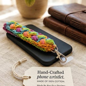 May include: A hand-crafted phone wristlet made of 100% cotton. The wristlet features a colorful, textured design with a silver-tone clasp. The wristlet is resting on a black phone case. The tag reads "Hand-Crafted phone wristlet. Made by Bee Ewe Designs."